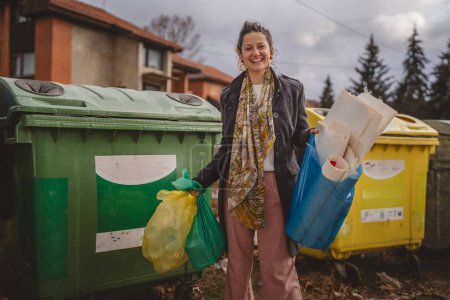 Téléchargez les photos : Un stand de femme adulte avec des sacs verts jaunes et bleus devant des conteneurs de recyclage pour le verre, le papier et le plastique zéro déchet concept écologique - en image libre de droit