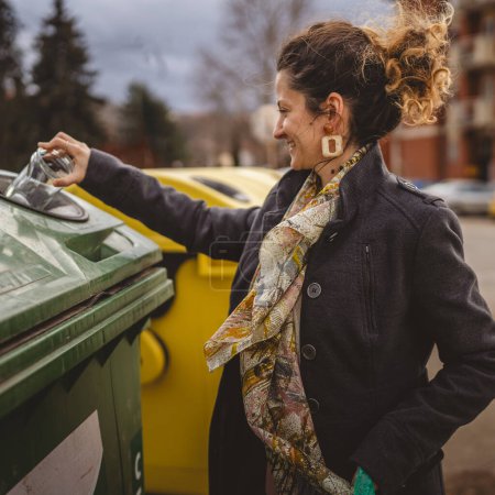Téléchargez les photos : Un stand de femme adulte avec des déchets de verre et des sacs verts devant des conteneurs de recyclage jetant un pot de bouteille pour réutiliser zéro déchet concept écologique - en image libre de droit