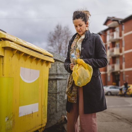 Foto de Una mujer adulta de pie con residuos de plástico y bolsas amarillas en frente de contenedores de reciclaje lanzando botella para la reutilización de residuos cero concepto ecológico - Imagen libre de derechos
