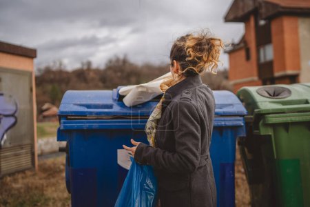 Foto de Una mujer adulta de pie con residuos de papel y bolsas azules en frente de contenedores de reciclaje lanzando para la reutilización de residuos cero concepto ecológico - Imagen libre de derechos