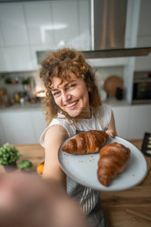 Photo for One adult caucasian woman stand in the kitchen with fresh croissant for breakfast daily morning routine - Royalty Free Image