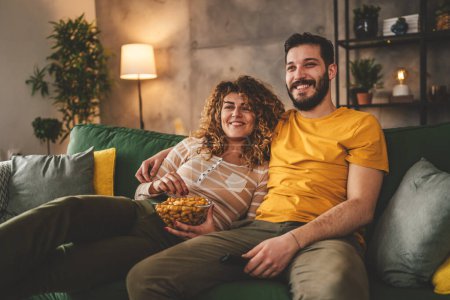 Téléchargez les photos : Couple homme et femme caucasien mari et femme ou petit ami et petite amie assis sur le canapé-lit à la maison regarder la série de films télévisés avec tenir télécommande heureux sourire lien amour famille concept - en image libre de droit