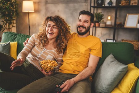 Téléchargez les photos : Couple homme et femme caucasien mari et femme ou petit ami et petite amie assis sur le canapé-lit à la maison regarder la série de films télévisés avec tenir télécommande heureux sourire lien amour famille concept - en image libre de droit