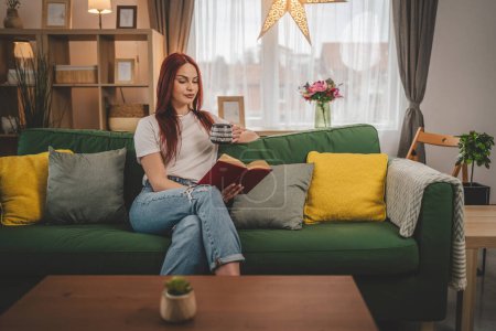 Téléchargez les photos : Jeune femme adulte une étudiante lit un livre ou étudie à la maison espace de copie de cheveux rouges - en image libre de droit