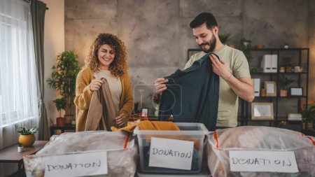 Photo for Husband and wife man and woman couple stand at home choose clothes for donation sorting wardrobe to the boxes in their apartment living room - Royalty Free Image