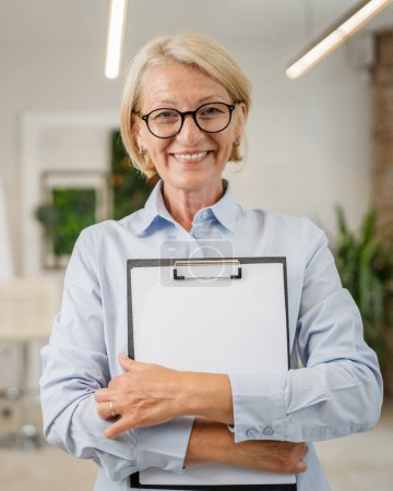 Téléchargez les photos : Portrait d'une femme caucasienne blonde mature avec des lunettes dans le bureau au travail sourire heureux regardant vers l'espace de copie confiant appareil photo - en image libre de droit