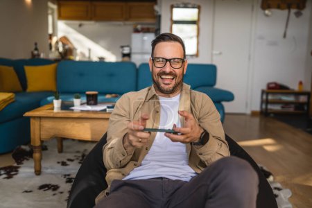 Photo for One adult caucasian man sit at home happy smile play video games leisure activity having fun hold mobile phone smartphone have fun copy space wear eyeglasses and shirt - Royalty Free Image
