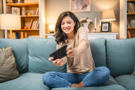 Téléchargez les photos : Une femme caucasienne adulte s'asseoir à la maison sourire heureux jouer à des jeux vidéo loisirs s'amuser tenir téléphone portable utiliser smartphone avoir du plaisir en ligne - en image libre de droit