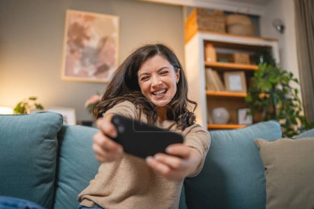 Téléchargez les photos : Une femme caucasienne adulte s'asseoir à la maison sourire heureux jouer à des jeux vidéo loisirs s'amuser tenir téléphone portable utiliser smartphone avoir du plaisir en ligne - en image libre de droit