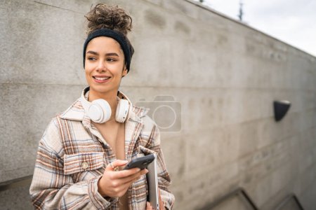 Photo for One woman young adult female student stand or walk outdoor hold laptop computer use smartphone phone for sms texting messages online real person copy space happy smile confident with headphones in day - Royalty Free Image