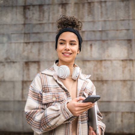 Foto de Una mujer joven adulta estudiante de pie o caminar al aire libre ordenador portátil uso teléfono inteligente para mensajes de texto SMS en línea persona real copiar espacio sonrisa feliz confiado con auriculares en el día - Imagen libre de derechos