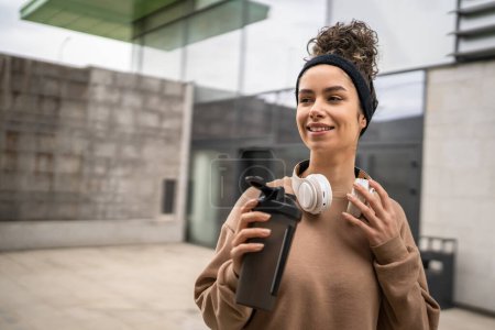 Photo for One young woman with headphones prepare for training hold supplement shaker bottle to drink water stand outdoor beautiful sporty caucasian female generation z healthy lifestyle concept copy space - Royalty Free Image