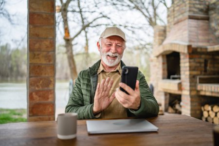 Téléchargez les photos : Un homme homme caucasien senior avec barbe grise et casquette assis en plein air au printemps ou en automne jour heureux sourire utiliser téléphone intelligent mobile pour l'espace de copie d'appel vidéo vraies personnes - en image libre de droit