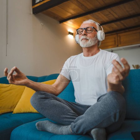 Foto de One man senior caucasian male eyes closed for guided training yoga or meditation while sitting at home with headphones self-care practice real people well-being inner peace and balance concept - Imagen libre de derechos