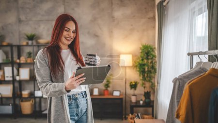 Téléchargez les photos : Femme stand à la fenêtre à la maison avec tablette numérique et tasse de café - en image libre de droit