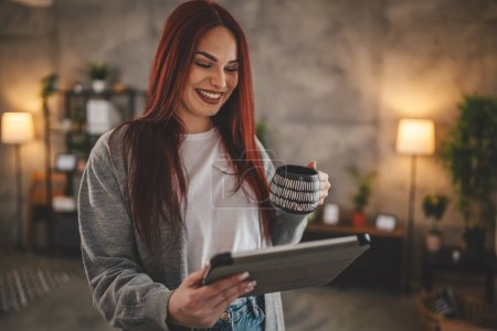 Téléchargez les photos : Femme stand à la fenêtre à la maison avec tablette numérique et tasse de café - en image libre de droit