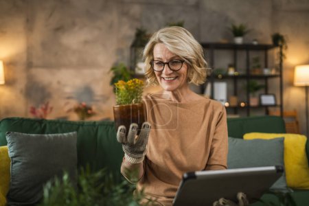 Téléchargez les photos : Une femme fleuriste prendre soin de cultiver des plantes à la maison tenir numérique tablette rechercher des conseils en ligne ou utiliser application application - en image libre de droit
