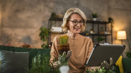 Téléchargez les photos : Une femme fleuriste prendre soin de cultiver des plantes à la maison tenir numérique tablette rechercher des conseils en ligne ou utiliser application application - en image libre de droit
