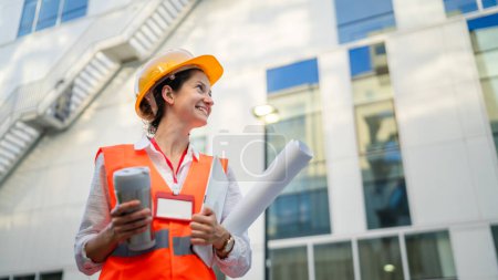 Téléchargez les photos : Architecte femme ingénieur de la construction debout à l'extérieur porter un casque de protection et à l'ouest en face du mur de bâtiment moderne - en image libre de droit