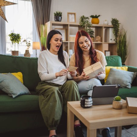 Foto de Dos mujeres adolescentes amigos o hermanas reciben regalos en la caja abierta tarjeta de lectura sonrisa feliz delante de la tableta digital en casa videollamada en línea - Imagen libre de derechos