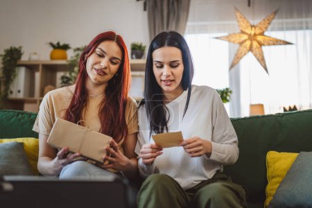 Foto de Dos mujeres adolescentes amigos o hermanas reciben regalos en la caja abierta tarjeta de lectura sonrisa feliz delante de la tableta digital en casa videollamada en línea - Imagen libre de derechos