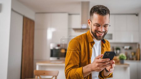 Téléchargez les photos : Homme avec des lunettes stand à la maison utilisation téléphone mobile sms textos - en image libre de droit
