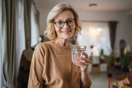 Téléchargez les photos : Gros plan portrait d'une femme mature avec un verre d'eau à la maison - en image libre de droit