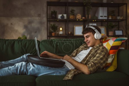 Foto de Un yong hombre caucásico adolescente masculino sentarse en casa con auriculares ver video de película en línea o serie en el ordenador portátil - Imagen libre de derechos
