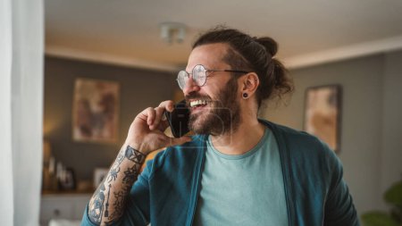 Téléchargez les photos : Un homme moderne adulte avec des lunettes de vue barbe et les cheveux longs stand à la maison utilisation téléphone mobile parler - en image libre de droit