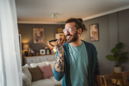 Téléchargez les photos : Un homme moderne adulte avec des lunettes de vue barbe et les cheveux longs stand à la maison utilisation téléphone mobile parler - en image libre de droit