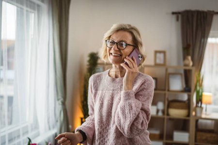 Téléchargez les photos : Une femme blonde mature debout à la maison utiliser téléphone mobile parler - en image libre de droit