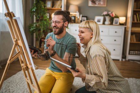 Téléchargez les photos : Couple peinture sur toile chevalet à la maison pinceau aider à apprendre à faire peinture pinceau tenir heureux homme et femme dans l'amour petit ami et petite amie ou mari et femme - en image libre de droit