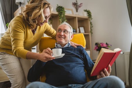 Téléchargez les photos : Homme âgé lire livre tandis que sa femme lui apporter une tasse de café à la maison - en image libre de droit