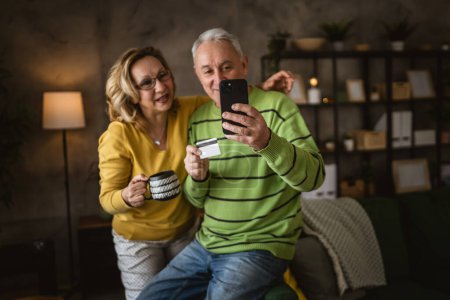 Téléchargez les photos : Couple mature homme caucasien senior et femme mari et femme à la maison shopping en ligne e-banking détenir carte de crédit ou de débit - en image libre de droit