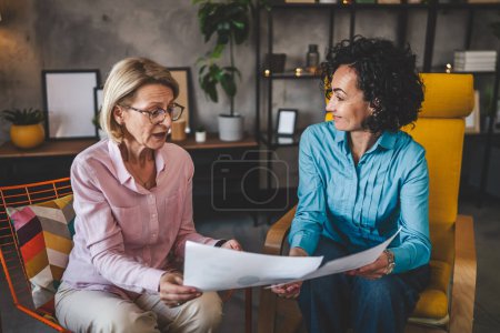 Photo for Two mature women sit at home hold paper document report have consultation businesswoman talk about actual tasks and problems - Royalty Free Image