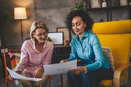 Photo for Two mature women sit at home hold paper document report have consultation businesswoman talk about actual tasks and problems - Royalty Free Image