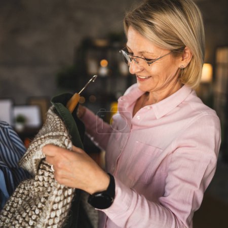 Foto de Una mujer rubia madura en casa elegir y ordenar la ropa en percha - Imagen libre de derechos