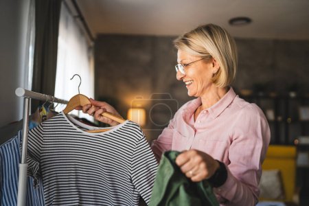 Photo for One mature blonde woman at home choose and sort clothes on hanger - Royalty Free Image