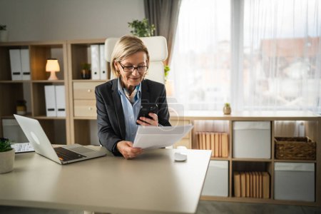 Ältere Frau blonde kausale Geschäftsfrau Unternehmerin oder Sekretärin sitzen im Büro Handy Smartphone verwenden, um Scan von Papier-Dokument oder Vertrag senden