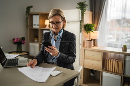 Ältere Frau blonde kausale Geschäftsfrau Unternehmerin oder Sekretärin sitzen im Büro Handy Smartphone verwenden, um Scan von Papier-Dokument oder Vertrag senden