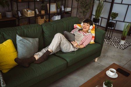 Téléchargez les photos : Femme Japonaise assise à la maison utiliser une tablette numérique pour regarder un film ou avoir un appel vidéo - en image libre de droit