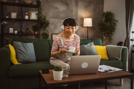 Téléchargez les photos : Les femmes japonaises tiennent une tasse de café et s'assoient avec un ordinateur portable à la maison prennent une pause du travail et regardent un film - en image libre de droit
