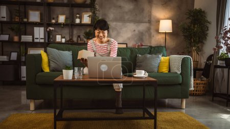 Téléchargez les photos : Une femme japonaise mature boîte ouverte présente et lit la carte lors d'un appel vidéo à la maison assis sur le canapé-lit - en image libre de droit