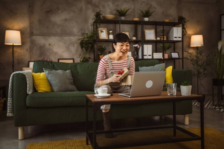 Téléchargez les photos : Une femme japonaise mature boîte ouverte présente et lit la carte lors d'un appel vidéo à la maison assis sur le canapé-lit - en image libre de droit