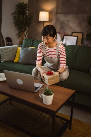 Foto de Una mujer japonesa madura caja abierta presenta y leer la tarjeta mientras en la videollamada en casa sentarse en el sofá cama - Imagen libre de derechos