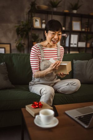 Foto de Una mujer japonesa madura caja abierta presenta y leer la tarjeta mientras en la videollamada en casa sentarse en el sofá cama - Imagen libre de derechos