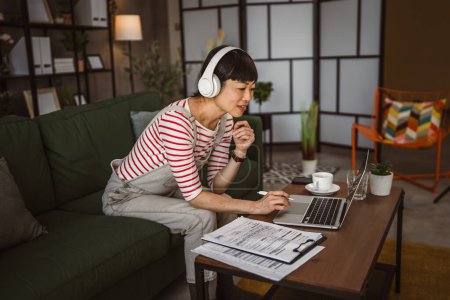 Téléchargez les photos : Femme japonaise travailler à la maison à l'ordinateur portable et détenir un document - en image libre de droit