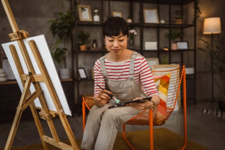 Téléchargez les photos : Une femme artiste peinture toile sur chevalet tenir pinceau à la maison - en image libre de droit