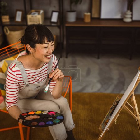 Téléchargez les photos : Une femme artiste peinture toile sur chevalet tenir pinceau à la maison - en image libre de droit