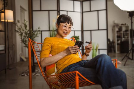 Téléchargez les photos : Japonais mature femme avec des lunettes assis sur chaise utiliser carte de crédit et téléphone mobile achats en ligne à la maison - en image libre de droit